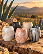 Cinco Vasos de mármol y ónix en tonos blancos, negros, grises y rosas, dispuestos sobre una superficie rocosa con un paisaje desértico al fondo.